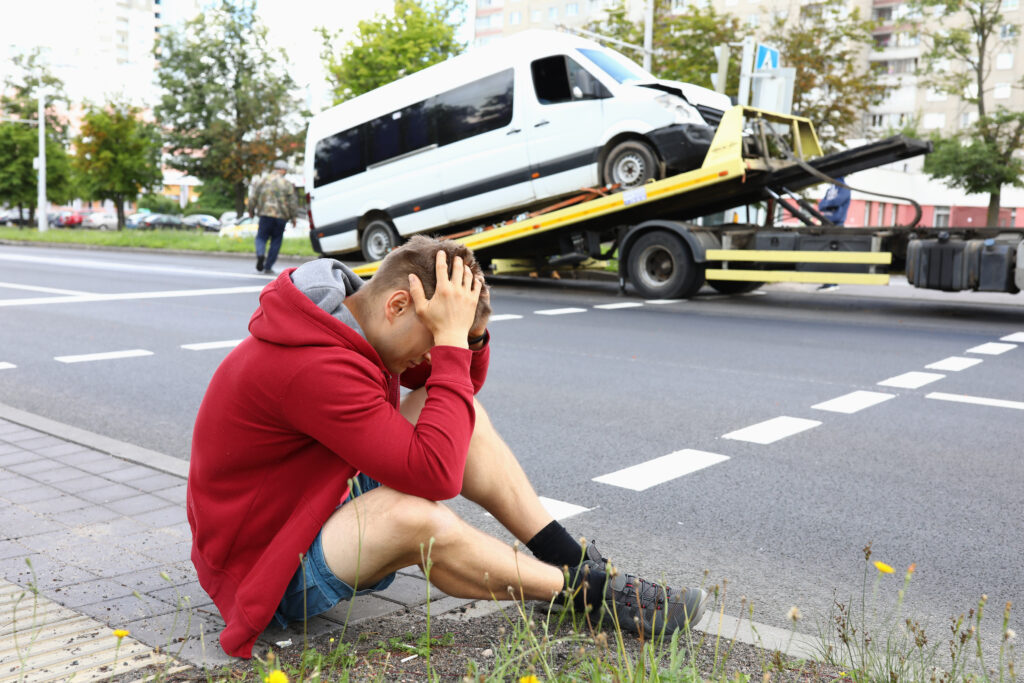 man sits on the sidewalk as mini van gets towed after a car accident